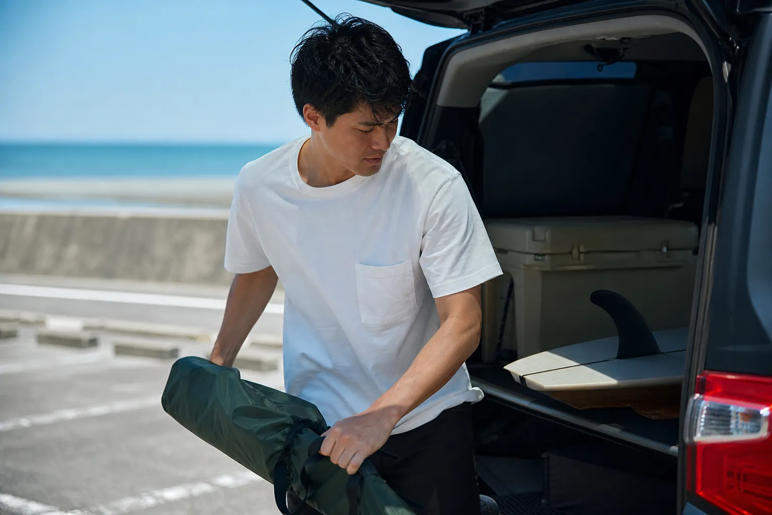 初夏の休日、海沿いの駐車場で、半袖Tシャツ姿の30代の日本人男性が車の後部からレジャー用品を降ろしている