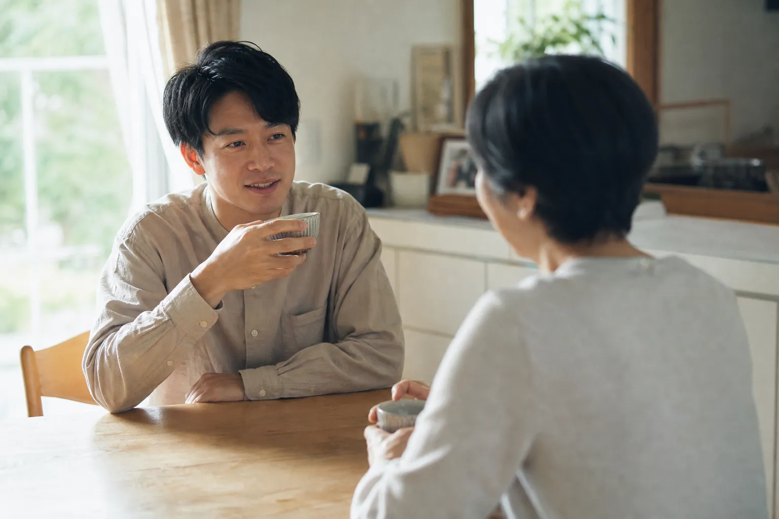 休日の昼、実家のダイニングテーブルで、30代の日本人男性が母親と向かい合って湯呑みを手に穏やかに話している