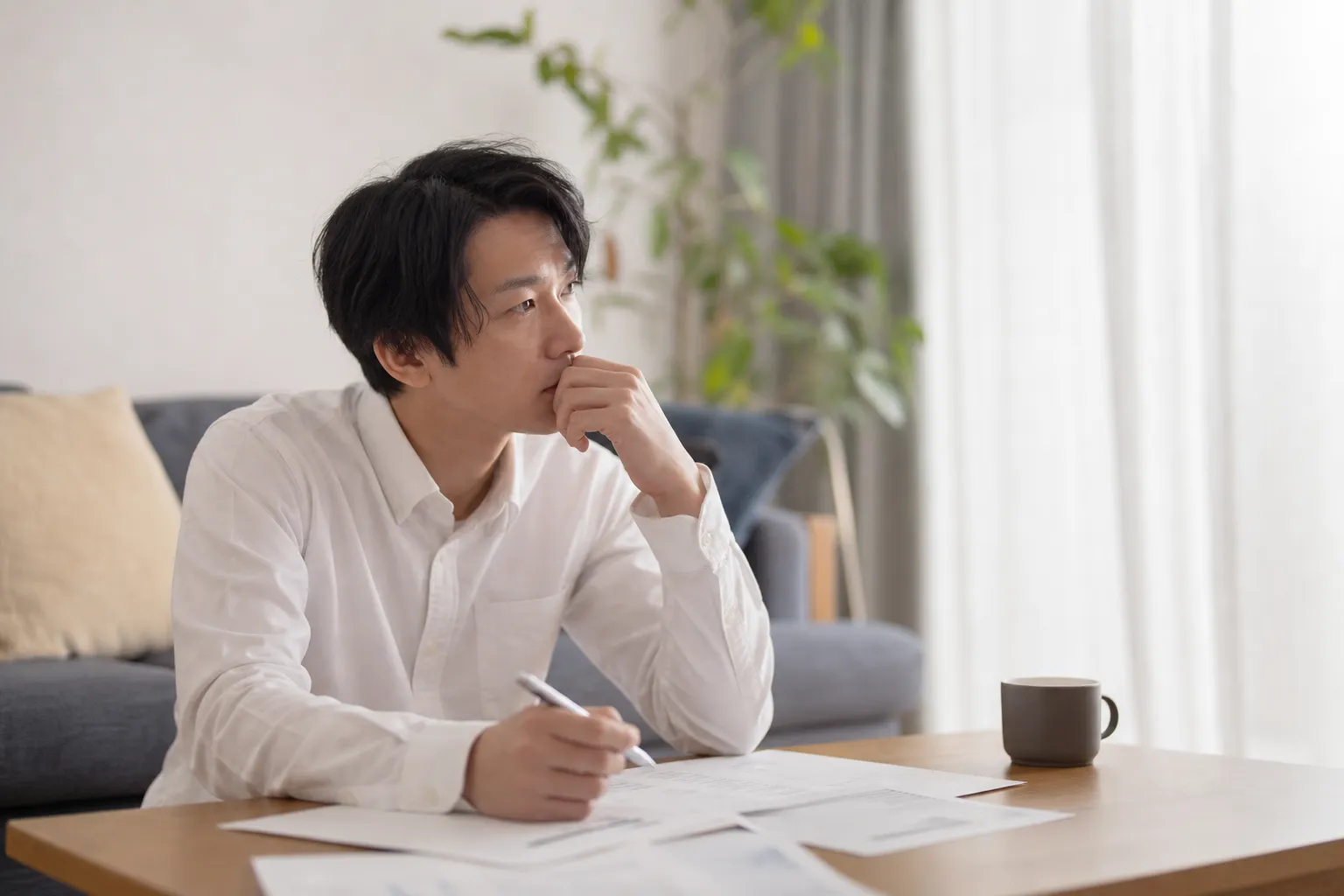 休日の午後、自宅のリビングのローテーブルに書類を広げ、30代の日本人男性がペンを持ったまま手を止めて考え込んでいる