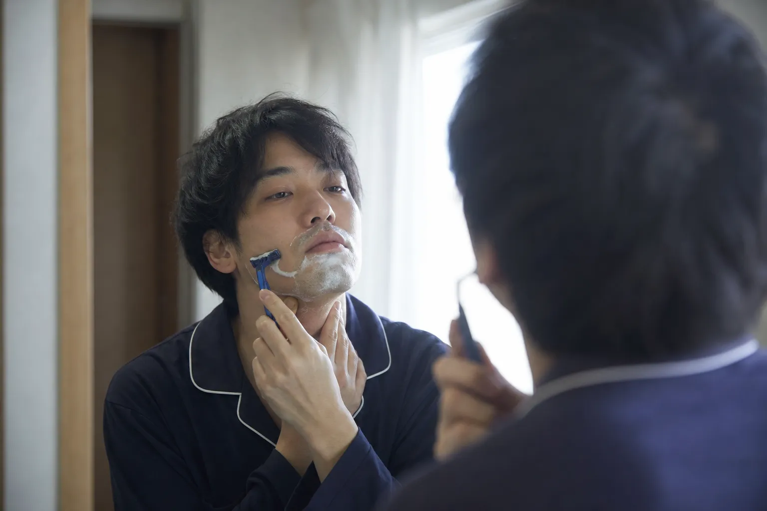 30代の日本人男性が早朝の洗面所で、鏡に向かってT字カミソリでヒゲを剃っている