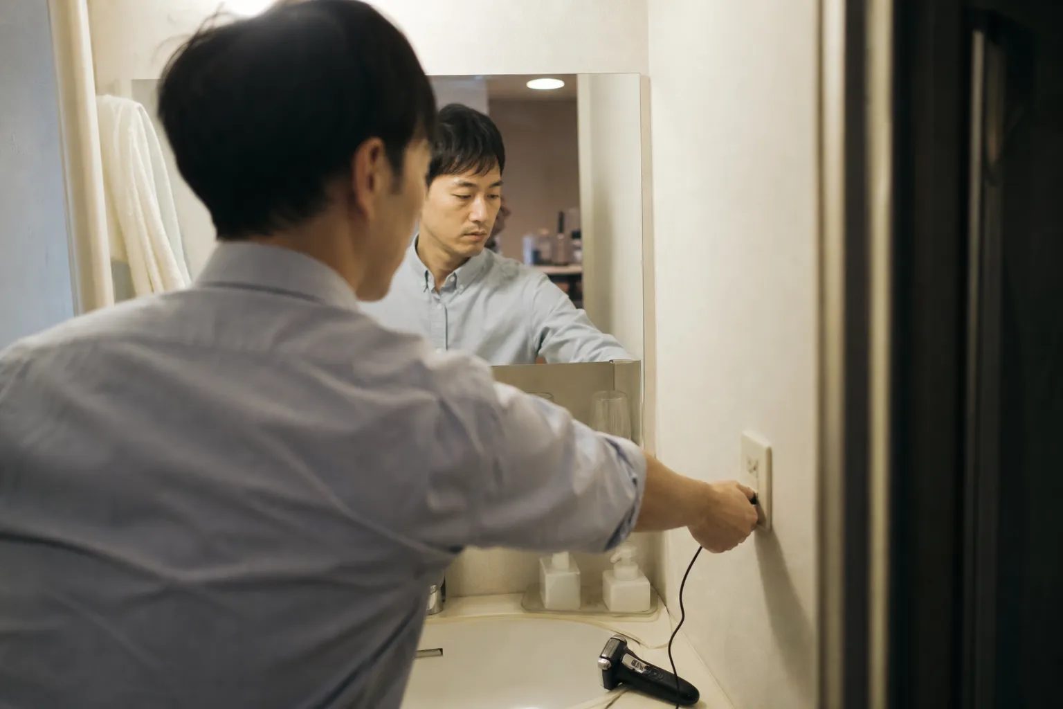 出張先のビジネスホテルの狭い洗面台で、30代の日本人男性が持参した電気シェーバーの充電コードをコンセントに差し込んでいる