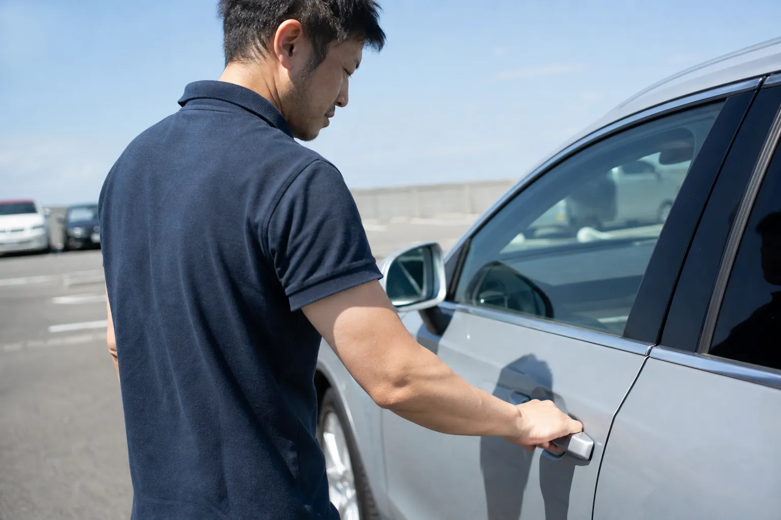 40代の日本人男性が真夏の昼、屋外の駐車場で車の運転席のドアを開けようとしている