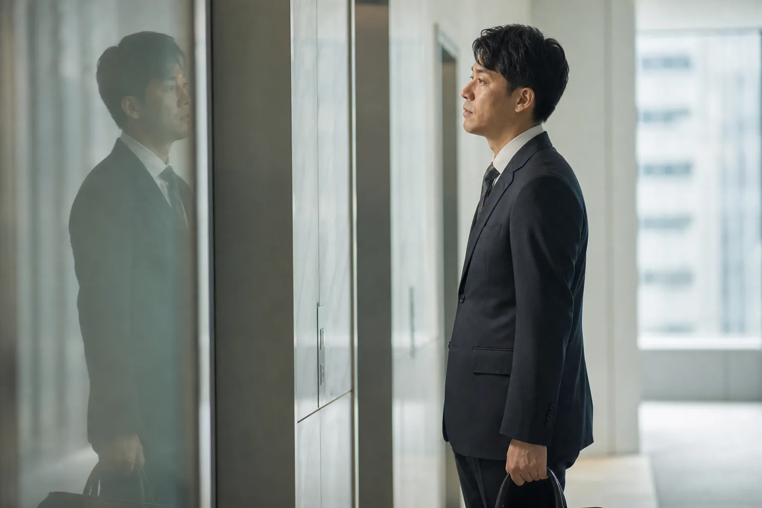 平日の朝、オフィスビルのエレベーターホールで、スーツ姿の30代の日本人男性がガラスに映った自分の顔をふと確かめている