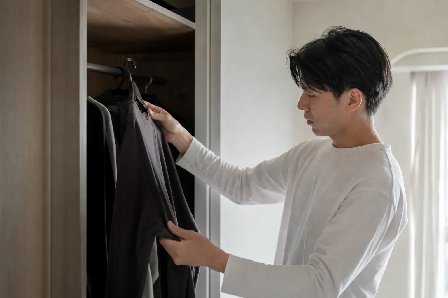 休日の朝、自宅のクローゼットの前で日本人の男性が、ハンガーに掛かった服を手に取りながら今日の服を選んでいる