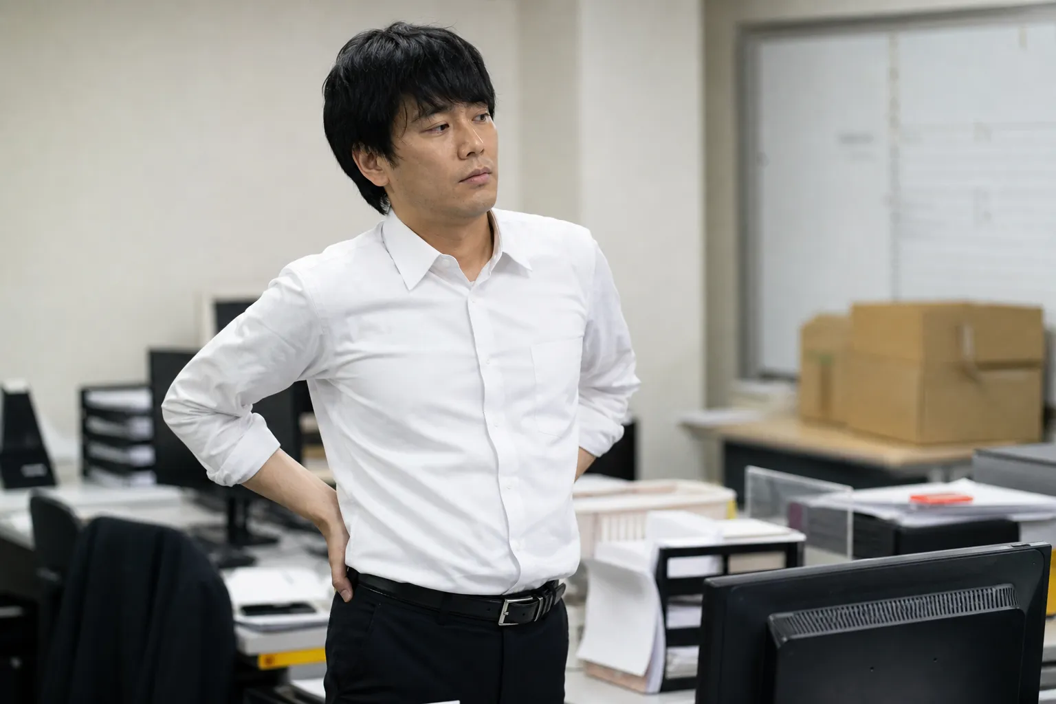 平日の夕方、オフィスのデスクで長時間座り続けた30代の日本人男性が、椅子から立ち上がって腰のあたりを軽くほぐしながら一息ついている