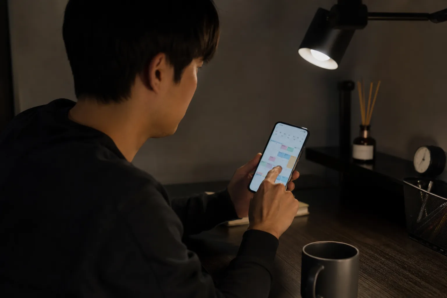 日本人の男性が自室の机でスマートフォンのカレンダーアプリを開き、3時間ほどの空き時間を指でなぞりながら予定を探している