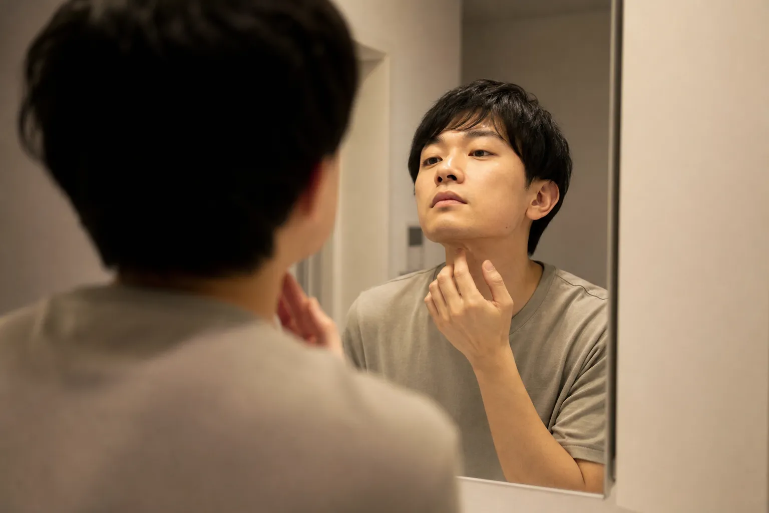 夜の自宅の洗面所で、日本人の男性が鏡に向かい、明日の準備としてあごの下を手で確かめている