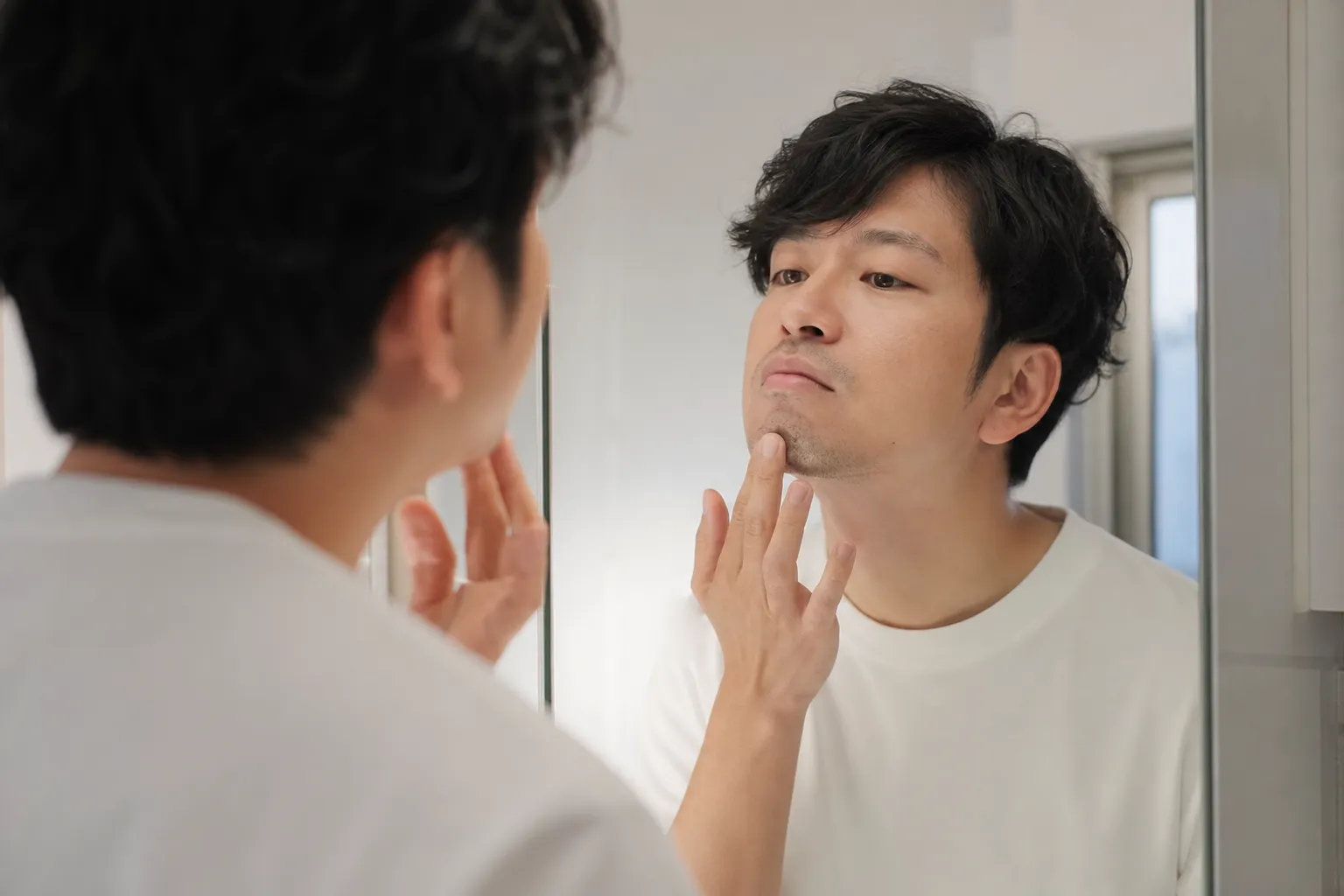 休日の昼、自宅の洗面所の鏡の前で、30代の日本人男性が顎のヒゲの剃り残しを指先で確かめている