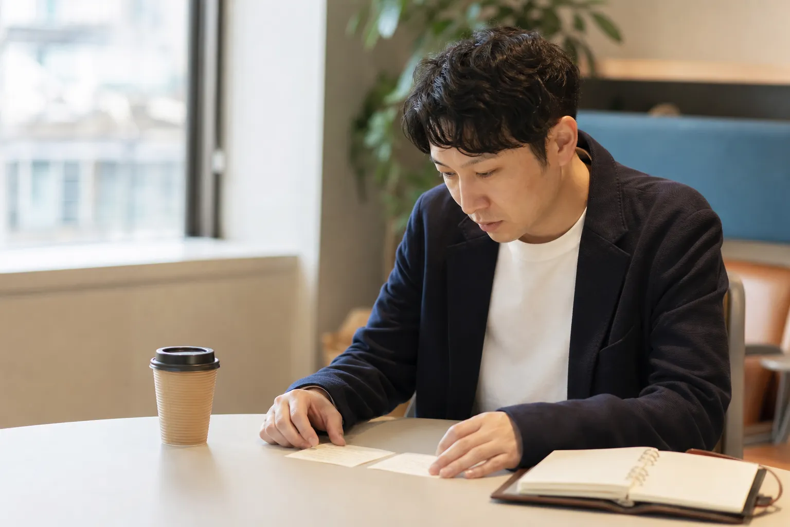 昼休みのオフィスの休憩スペースで、30代の日本人男性が紙コップのコーヒーを置いたまま、手帳に書いた2つのメモを並べて見比べている