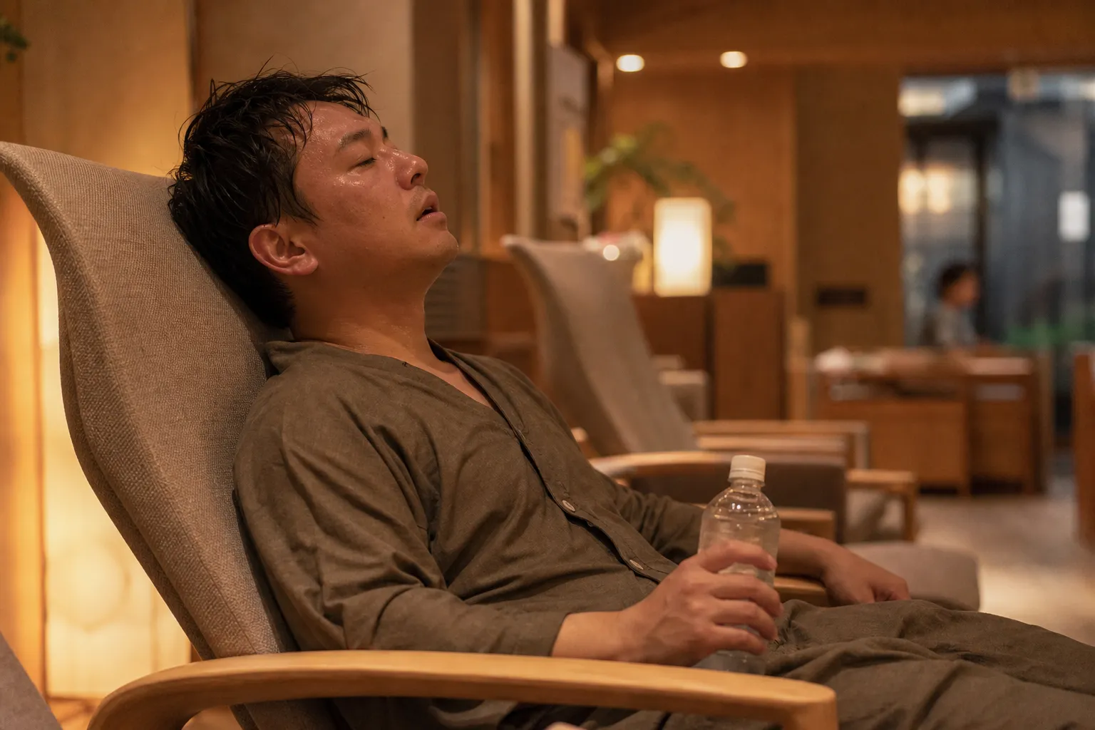 サウナ施設のロビーで、湯上がりの30代の日本人男性が館内着姿で椅子に深くもたれ、水のボトルを片手にほてった顔で息をついている