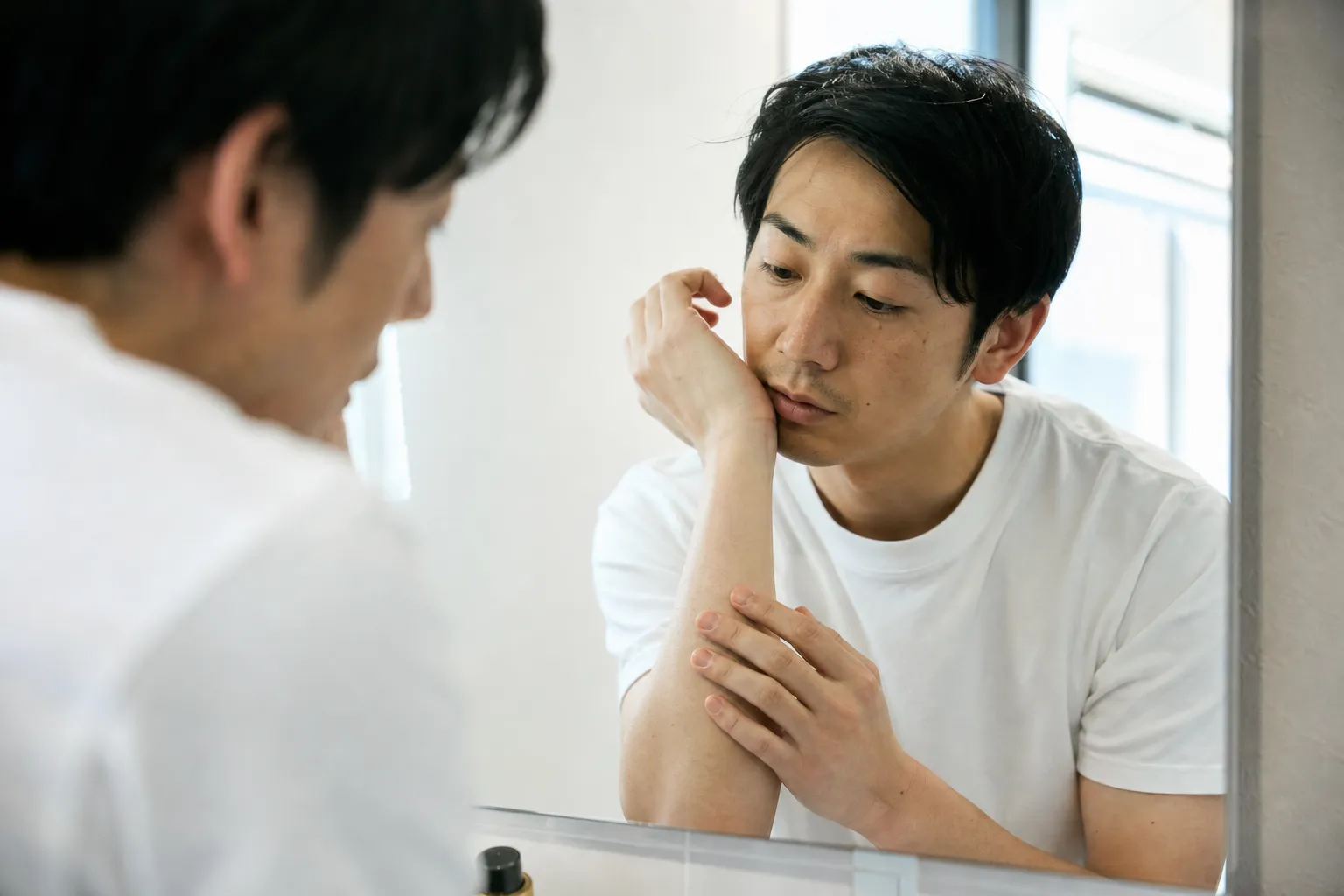 朝の洗面所で、鏡に腕を近づけて肌の様子をじっと確かめている30代の日本人男性