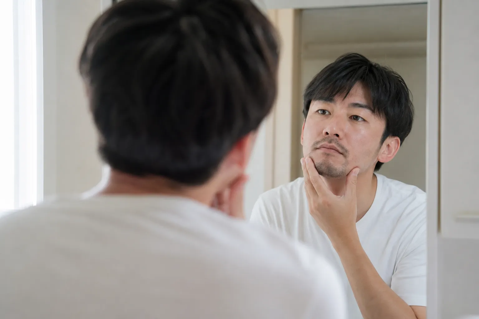 平日の朝、自宅の洗面所の鏡の前で、30代の日本人男性が片手をあごに当て、伸びかけのヒゲを確かめるように顔を少し傾けている