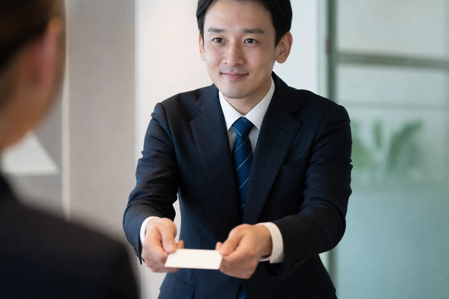 オフィスの会議室で、30代の日本人男性がスーツ姿で両手を添えて名刺を差し出しており、その手元がはっきり見えている